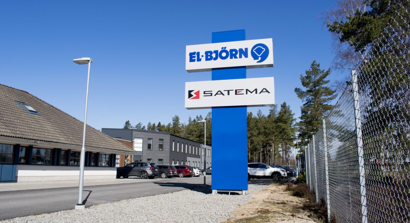 Fristående stolpskylt med ljuslådor för EL Björn och SATEMA, med EL Björns grå fastighet, tallar och blå himmel i bakgrunden