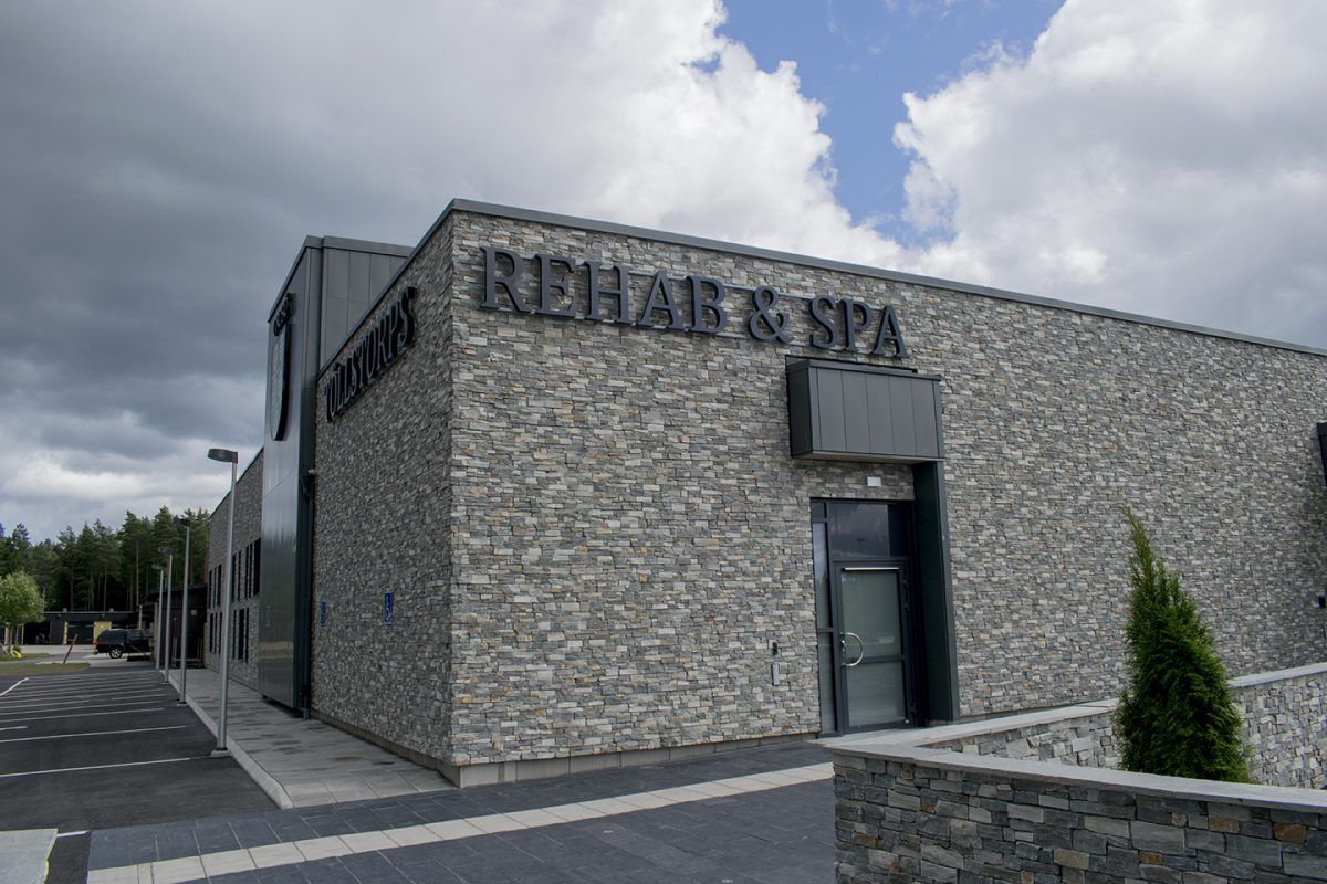 Vägg av natursten med bakåtlysande svart skylt med texten 'REHAB & SPA', vilket skapar en sofistikerad och inbjudande atmosfär