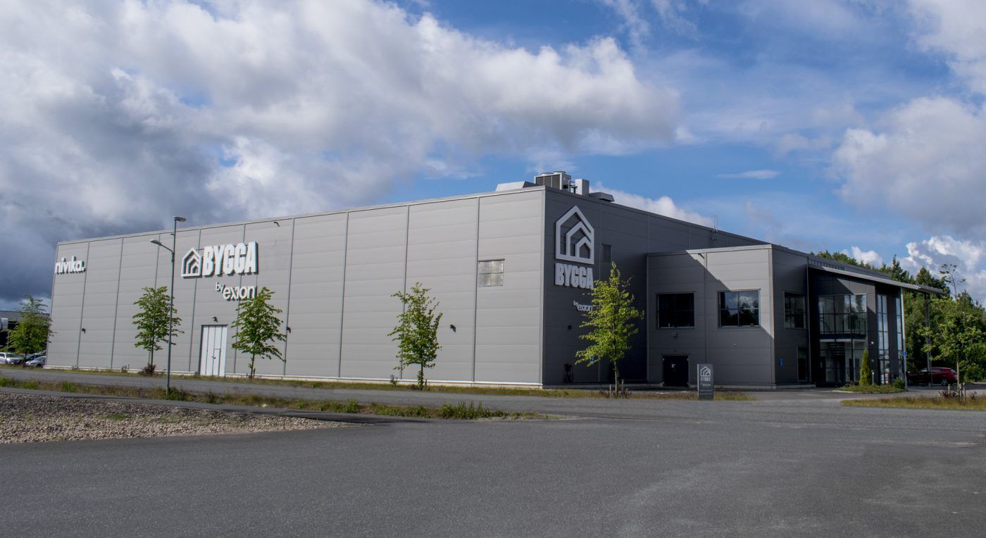Stor grå fastighet med vita fasadskyltar för BYGGA by exoion och Nivika, omgiven av blå himmel och vita moln, med asfalterad väg och gräsmatta i förgrunden