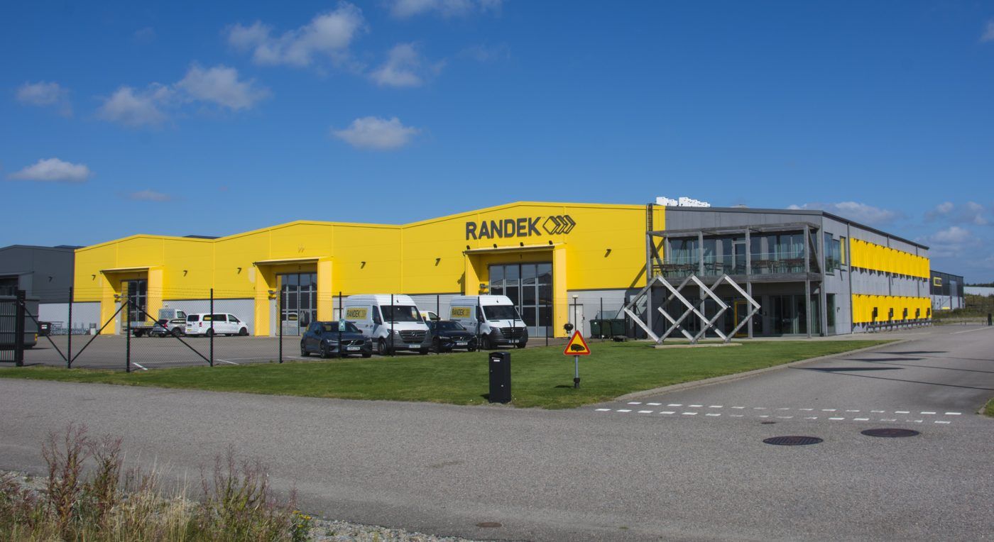 Stor gul och grå fastighet med företagsskylten 'RANDEK', omgiven av gräsmatta och parkering, med en blå himmel i bakgrunden