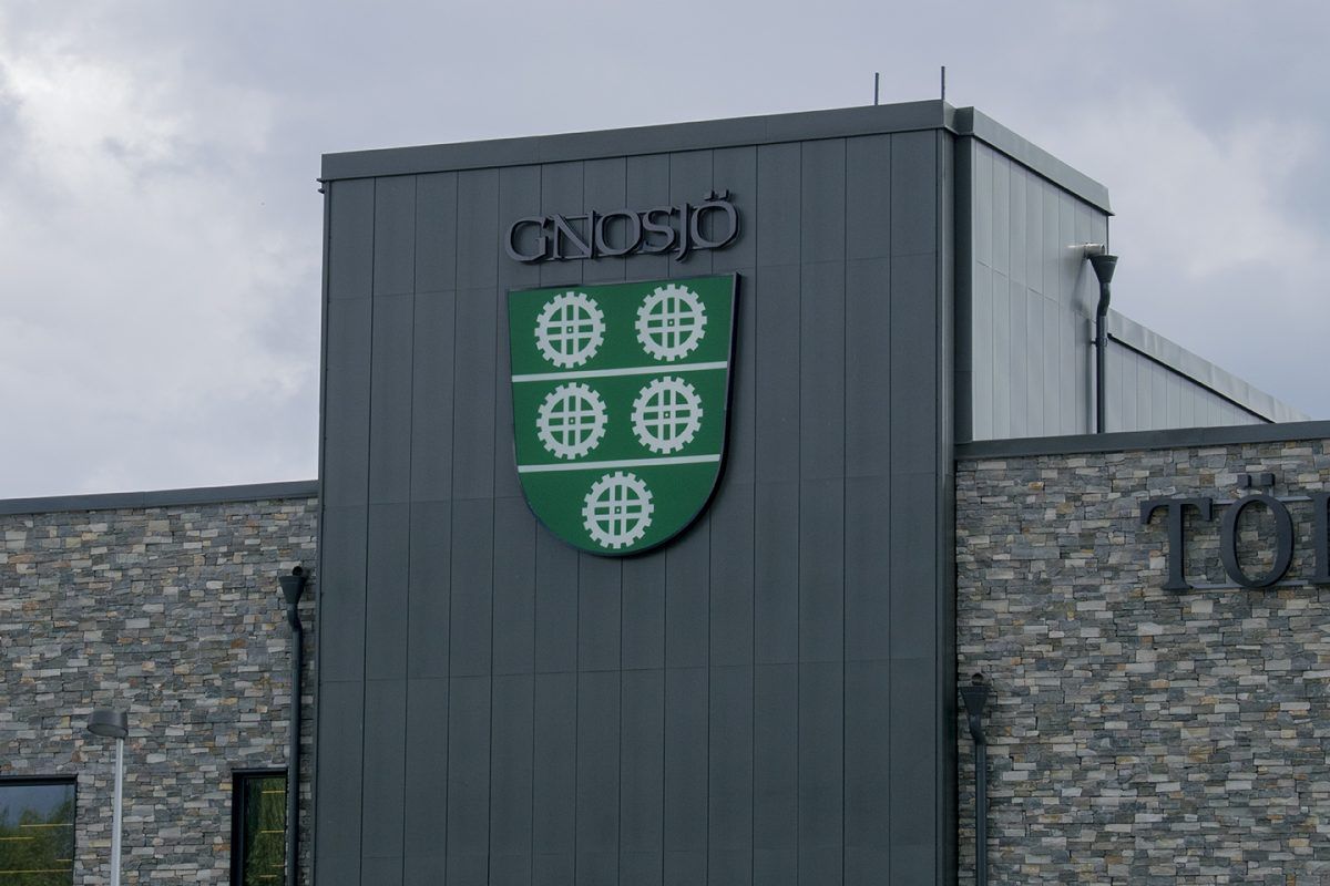 Grå träpanel med bakåtlysande texten 'Gnosjö' och en stor logotypsköld under, som ger en stilren och professionell visuell identitet