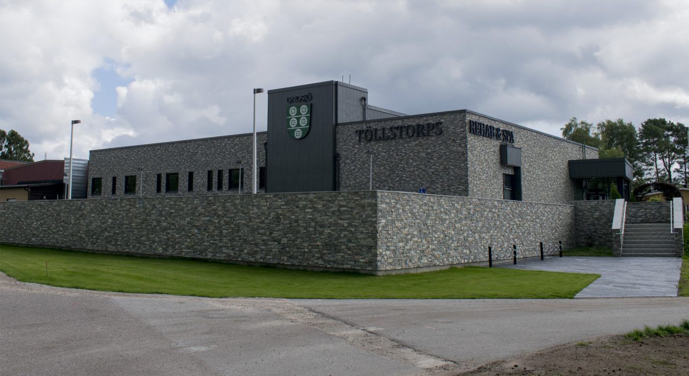 Töllstorp Rehab & Spa med byggnad i natursten och svarta bakåtlysande LED-bokstäver. En gråmålad träpanel pryds av texten Gnosjö och en stor sköld som en del av logotypen. Mur av natursten och blå himmel i bakgrunden