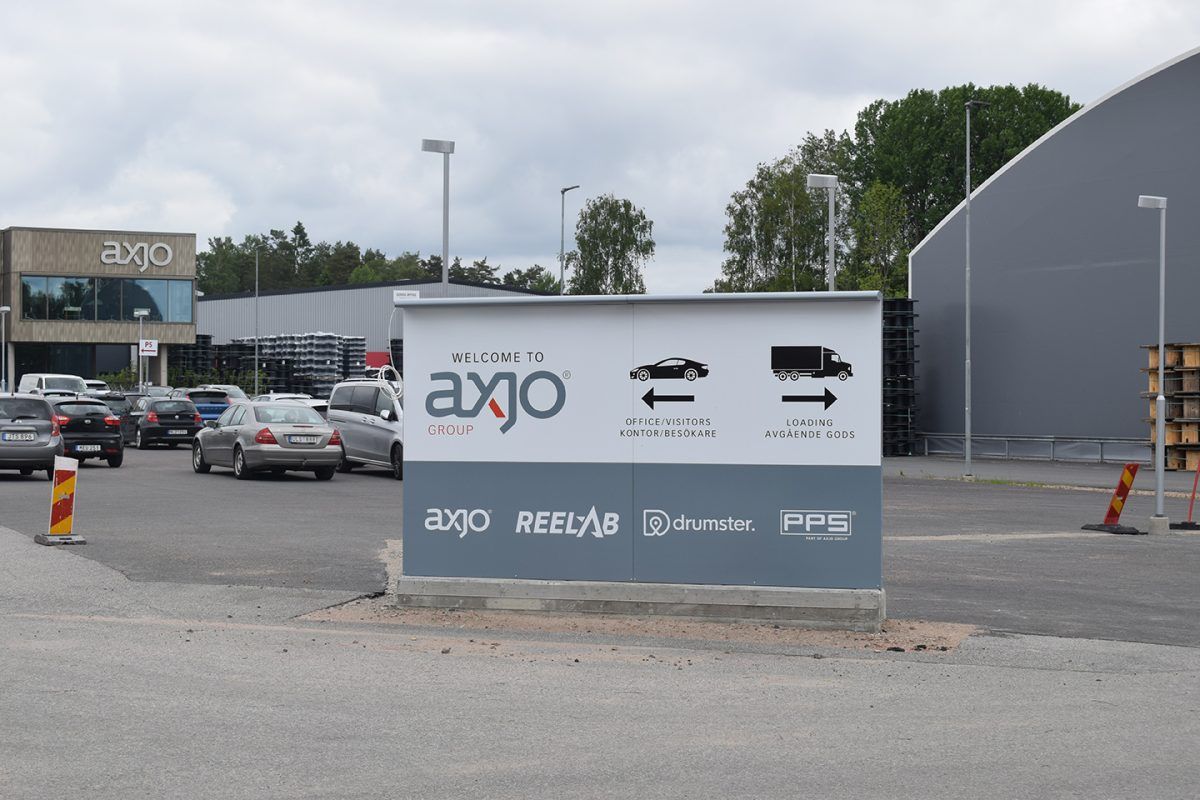 Fristående markskylt för AXJO med vita vägvisande pilar, som visar vänstersväng till kontoret och högersväng till godsmottagningen. Skylten är tillverkad i lackerad aluminium och placerad vid företagets infart