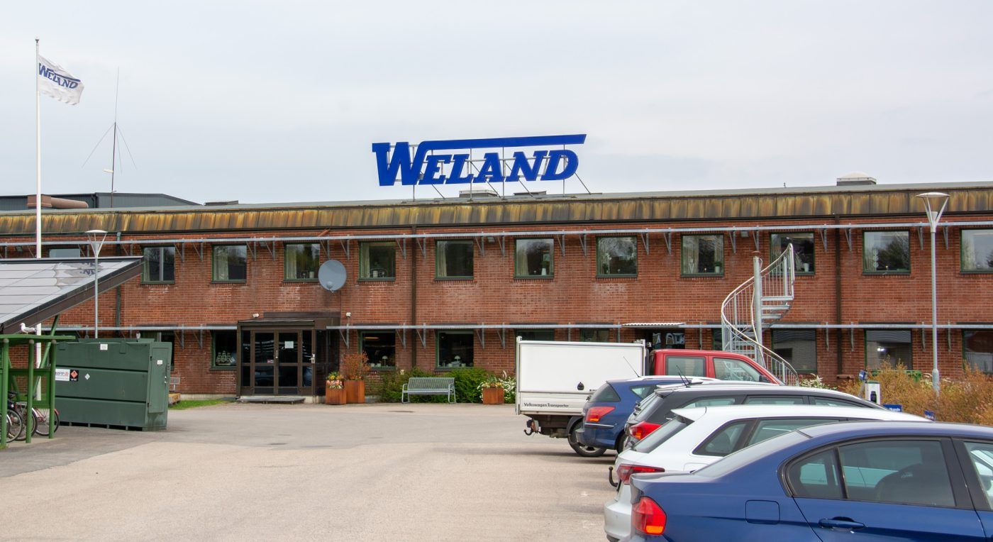 Weland AB:s stora tegelbyggnad med en takskylt från EpicSign, parkerade bilar i förgrunden och himmel i bakgrunden.