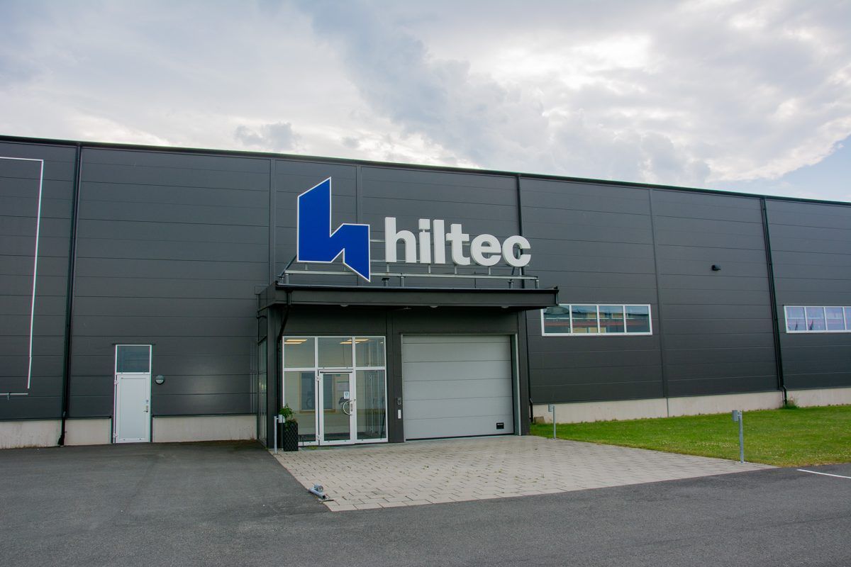 Hiltecs blå symbol och vita bokstäver ovanför entrétaket på en grå industribyggnad.
